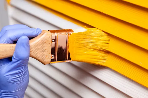 Gloved hand brushing yellow paint onto siding